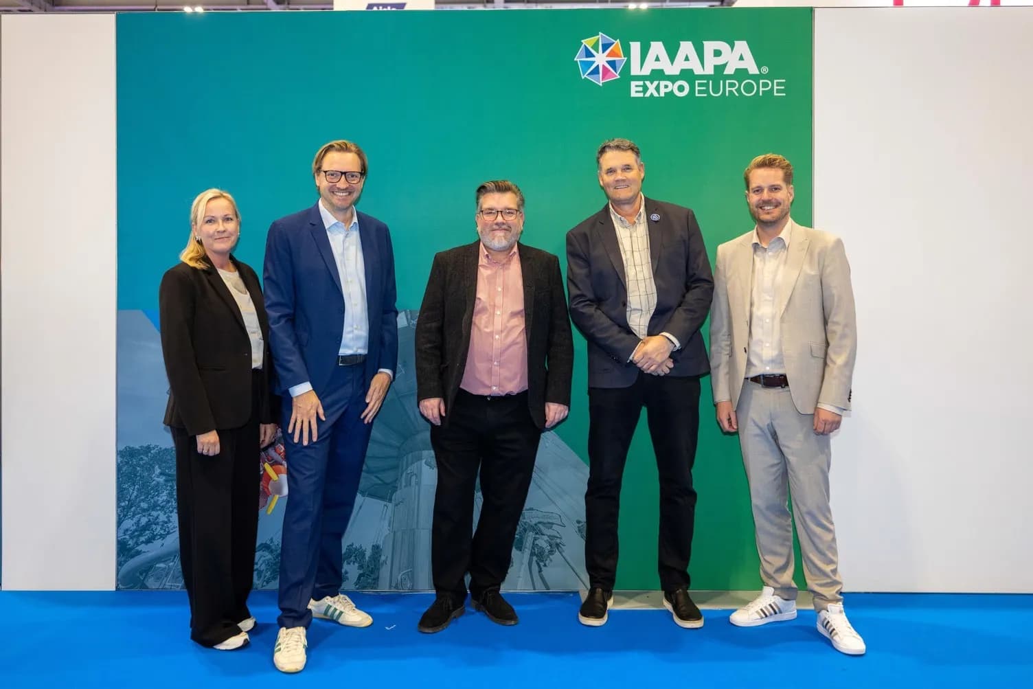 Five people in business attire pose for a photo in front of a green wall with the IAAPA Expo Europe logo.
